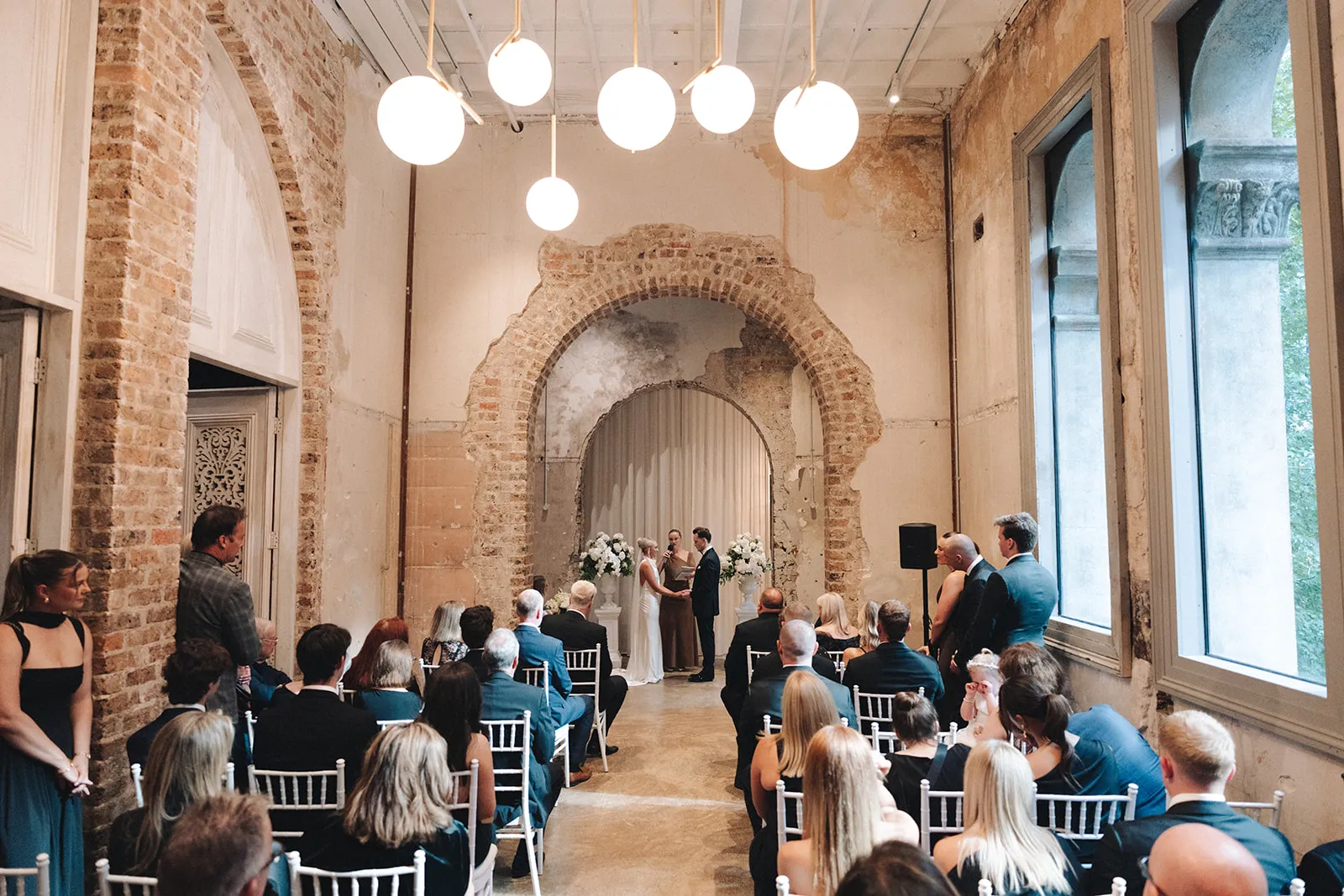 Guests seated for a ceremony in a heritage brick hall with globe lights